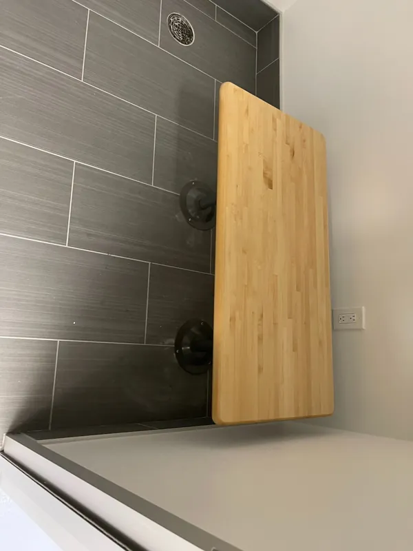 A folding wood shower seat mounted on a tiled shower wall